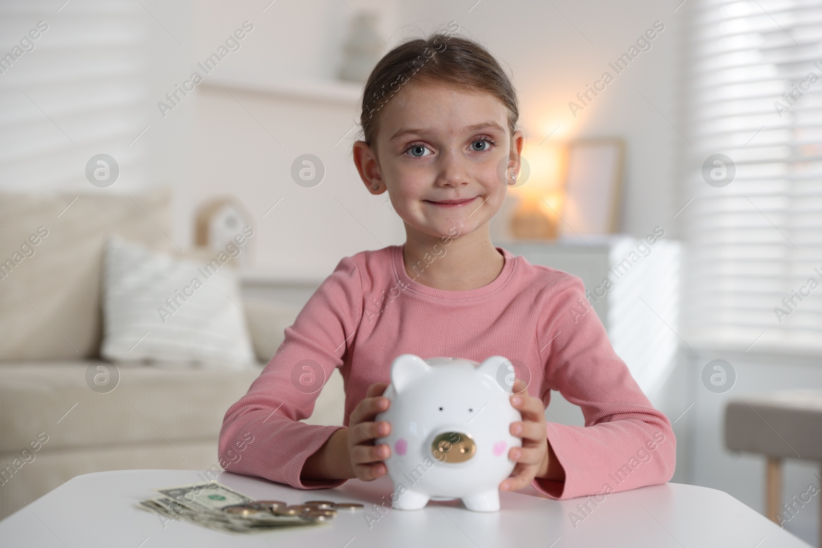 Photo of Little girl with pocket money and piggy bank at white table indoors