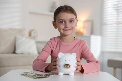 Photo of Little girl with pocket money and piggy bank at white table indoors