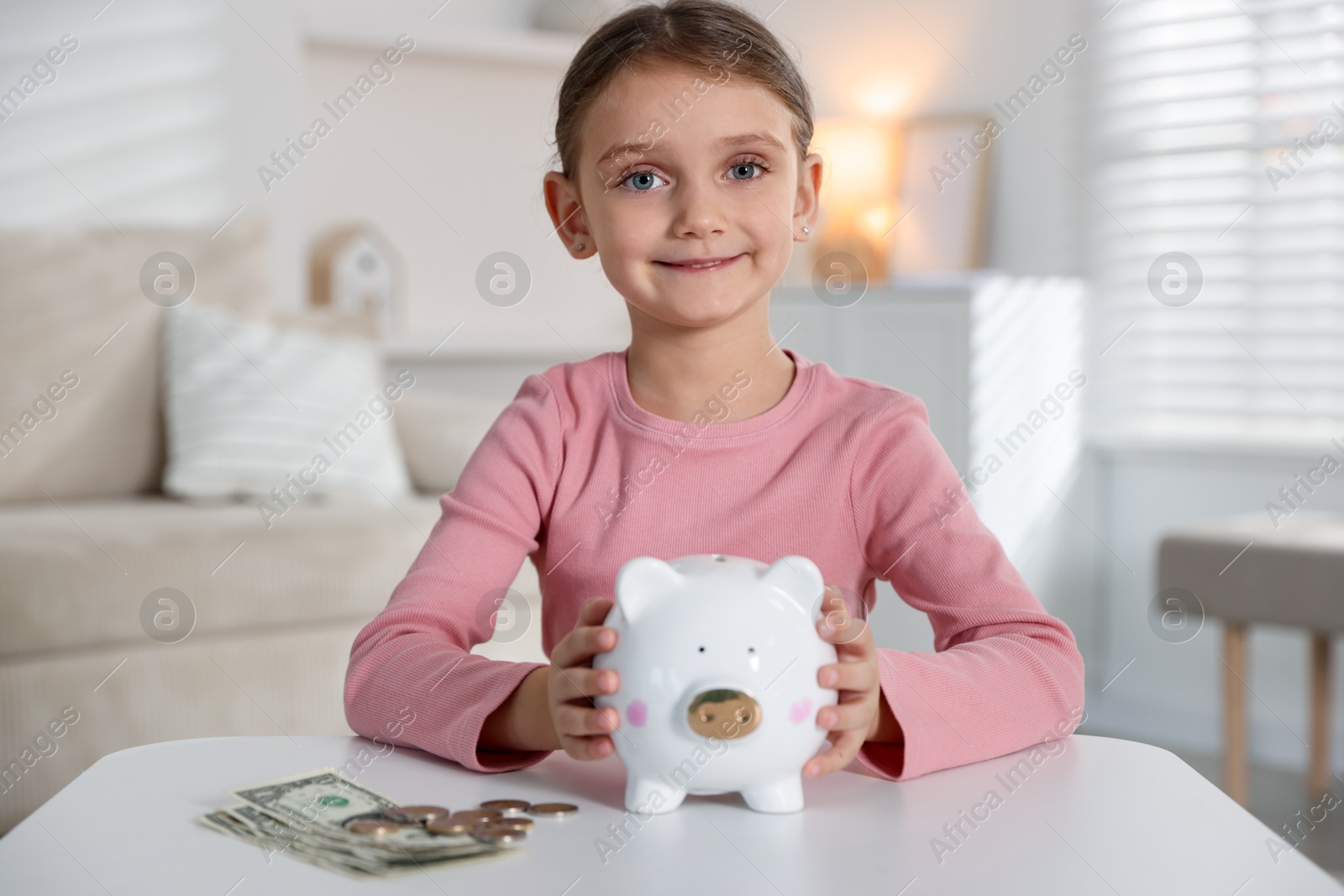 Photo of Little girl with pocket money and piggy bank at white table indoors