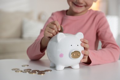 Photo of Little girl putting pocket money into piggy bank at white table indoors, closeup