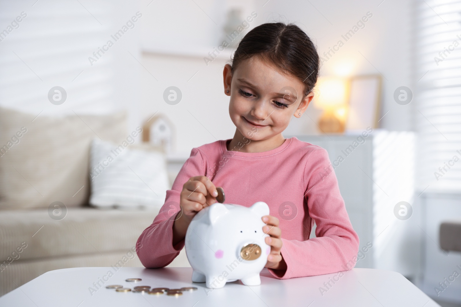 Photo of Little girl putting pocket money into piggy bank at white table indoors