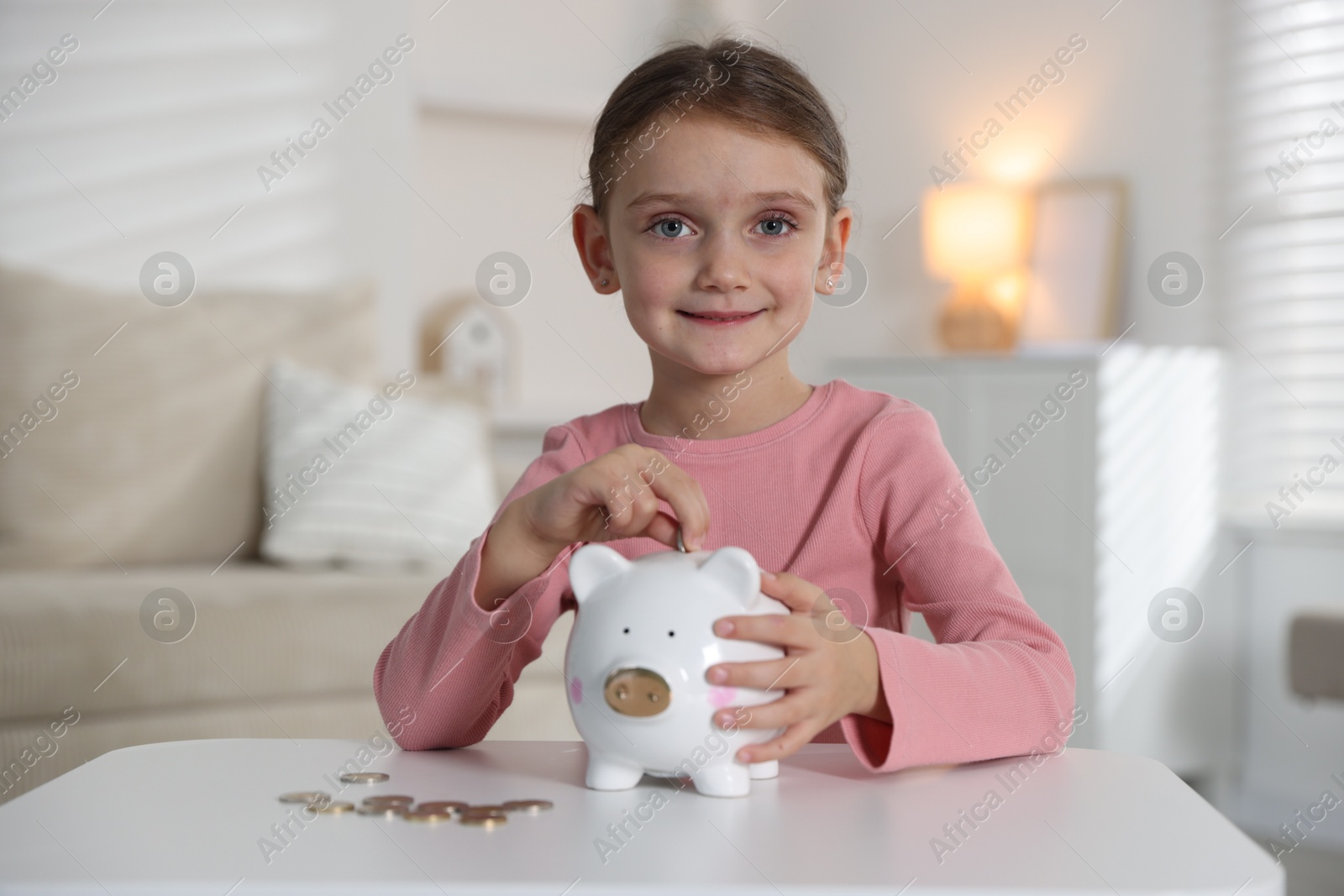 Photo of Little girl putting pocket money into piggy bank at white table indoors
