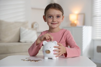 Photo of Little girl putting pocket money into piggy bank at white table indoors