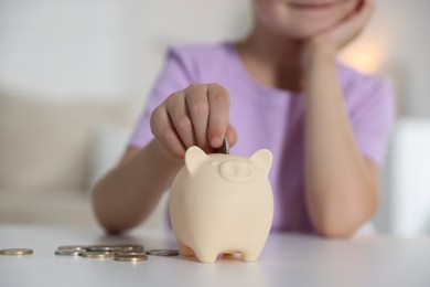 Photo of Little girl putting pocket money into piggy bank at white table indoors, closeup