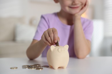 Photo of Little girl putting pocket money into piggy bank at white table indoors, closeup