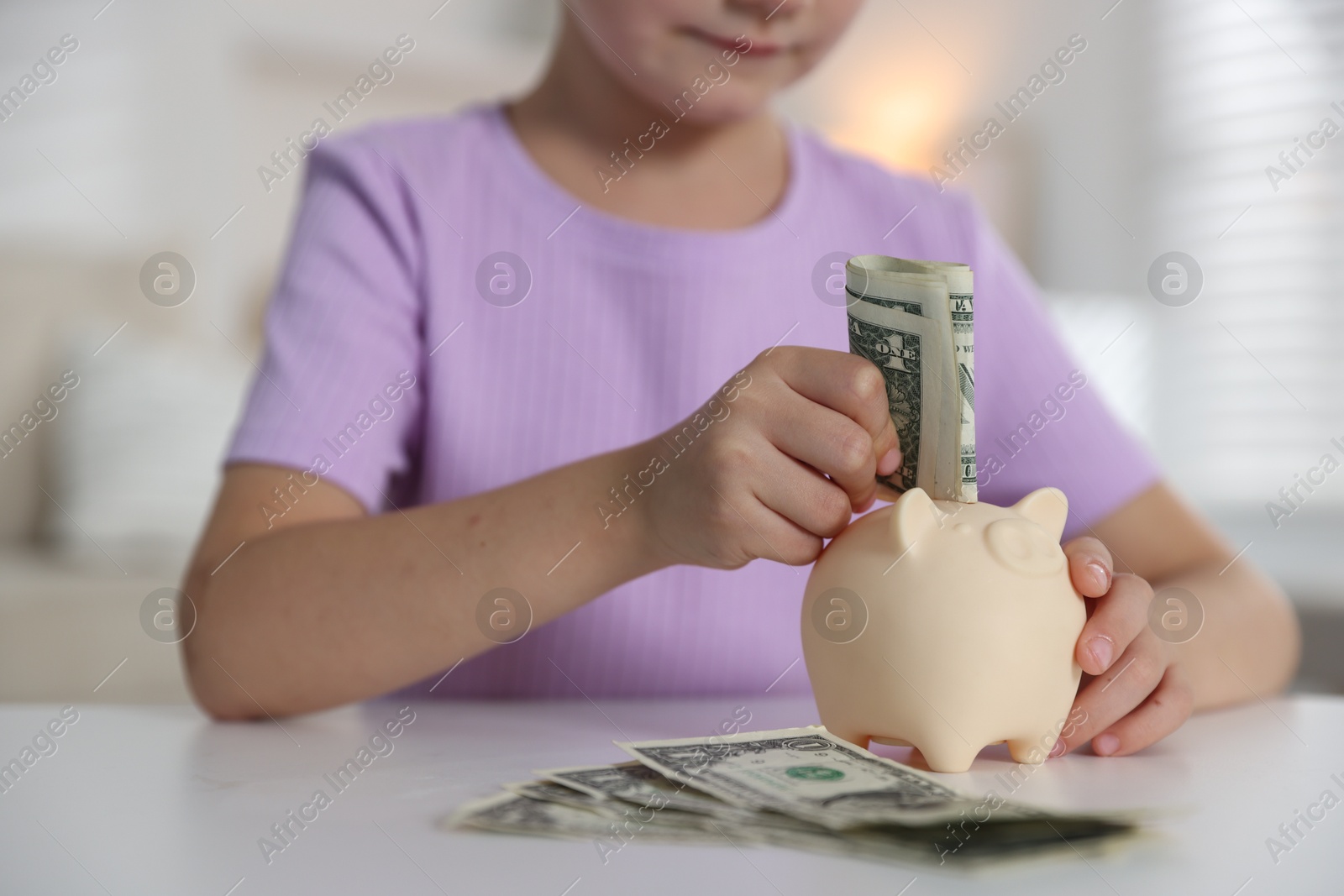 Photo of Little girl putting pocket money into piggy bank at white table indoors, closeup