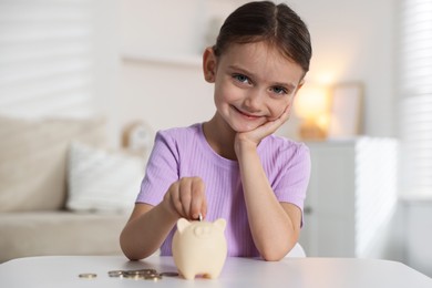 Photo of Little girl putting pocket money into piggy bank at white table indoors