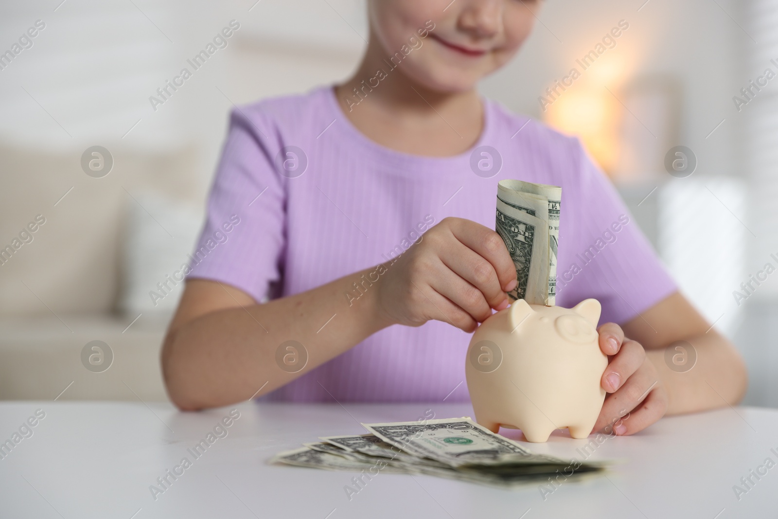 Photo of Little girl putting pocket money into piggy bank at white table indoors, closeup