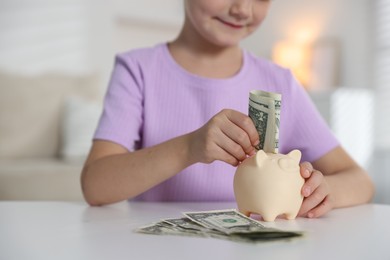 Photo of Little girl putting pocket money into piggy bank at white table indoors, closeup