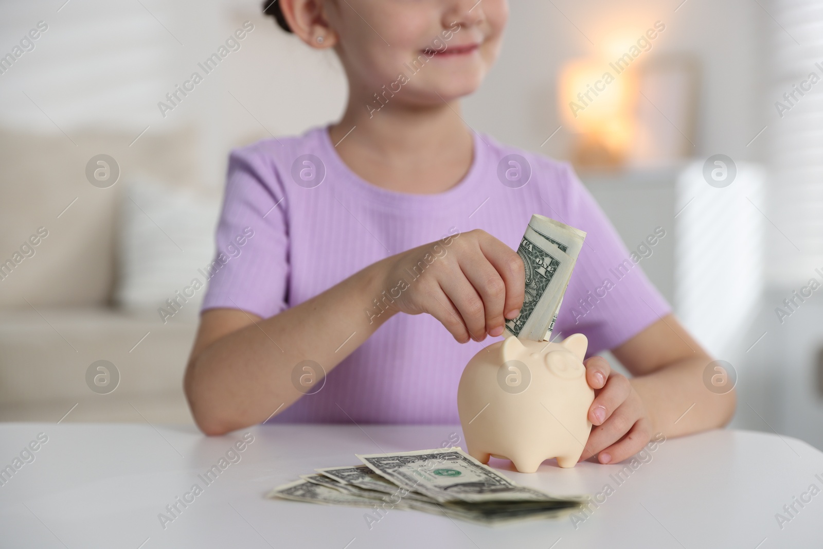Photo of Little girl putting pocket money into piggy bank at white table indoors, closeup