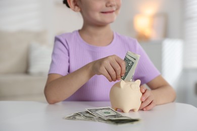 Photo of Little girl putting pocket money into piggy bank at white table indoors, closeup