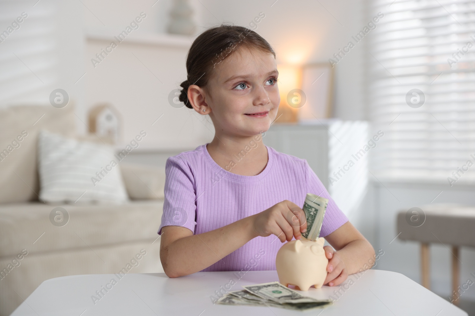Photo of Little girl putting pocket money into piggy bank at white table indoors