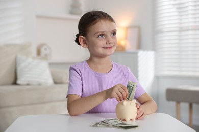 Photo of Little girl putting pocket money into piggy bank at white table indoors