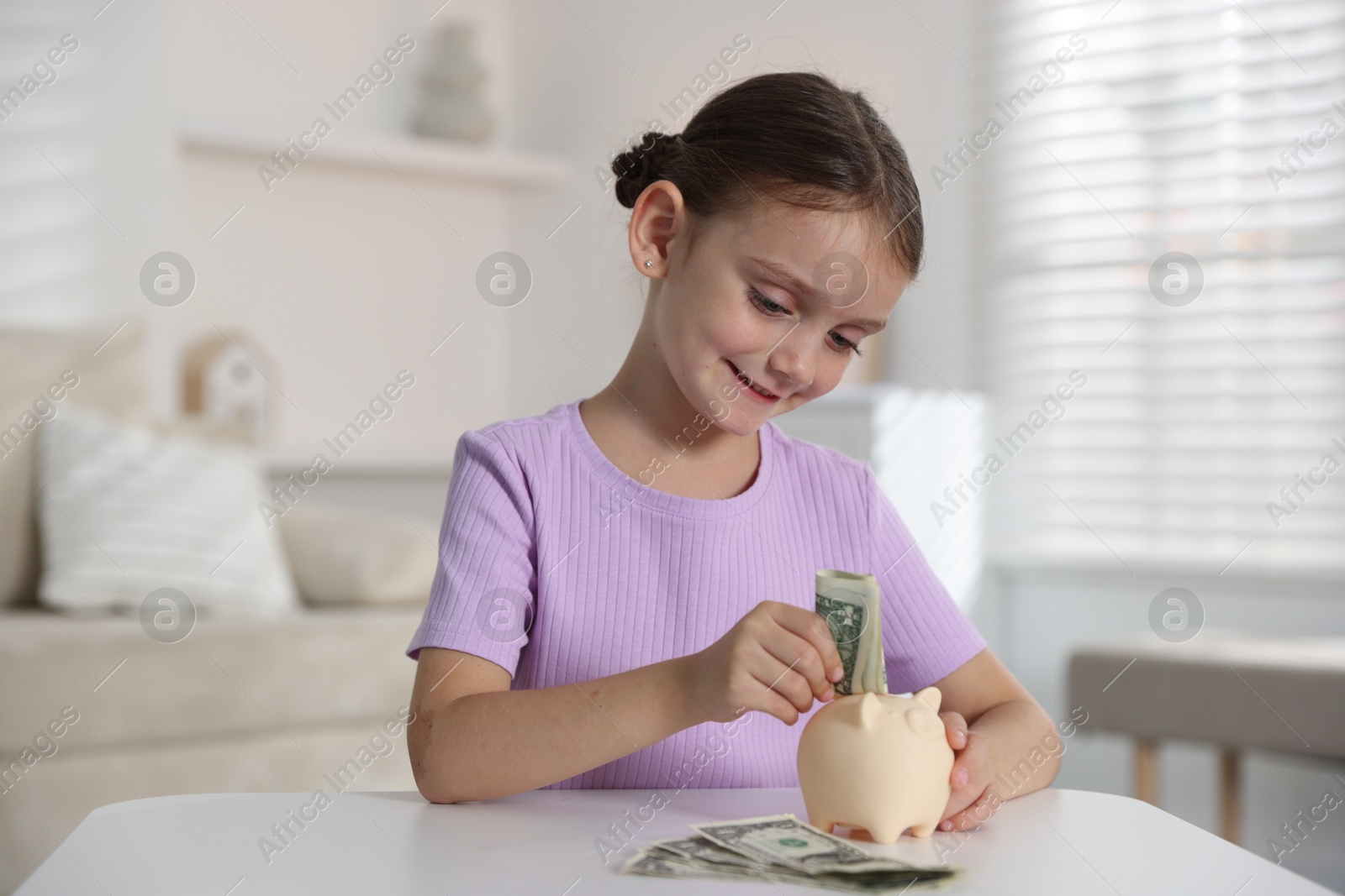 Photo of Little girl putting pocket money into piggy bank at white table indoors