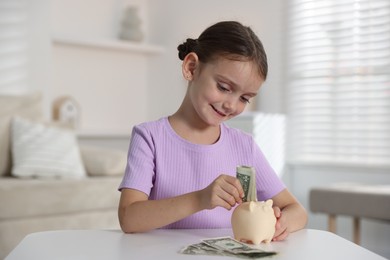 Photo of Little girl putting pocket money into piggy bank at white table indoors