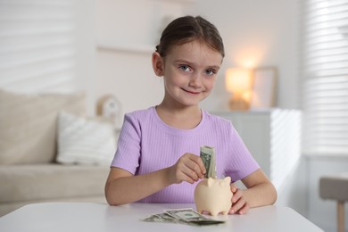 Photo of Little girl putting pocket money into piggy bank at white table indoors