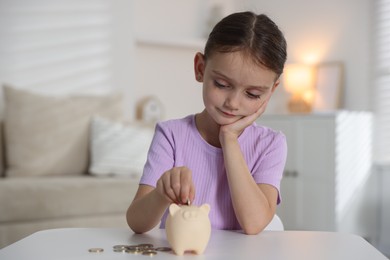 Photo of Little girl putting pocket money into piggy bank at white table indoors