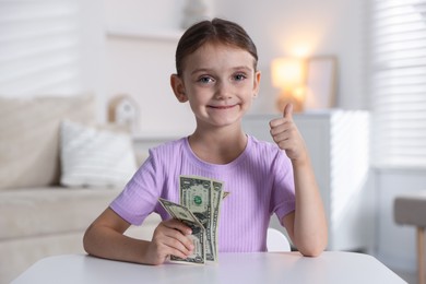 Photo of Little girl holding pocket money and showing thumbs up at white table indoors