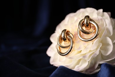 Photo of Stylish gold earrings and flower on blue silk cloth, closeup. Space for text