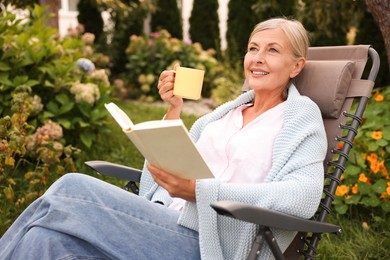 Photo of Senior woman with cup of drink reading book in armchair outdoors