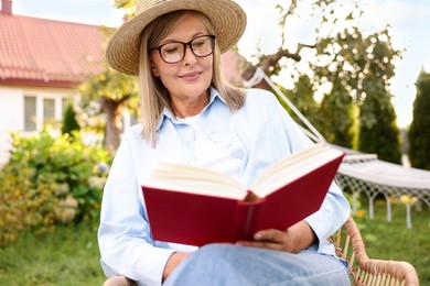 Photo of Senior woman reading book in wicker armchair outdoors