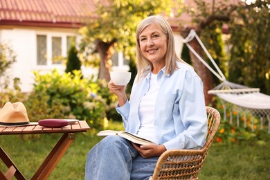 Photo of Senior woman with cup of drink and book in wicker armchair outdoors