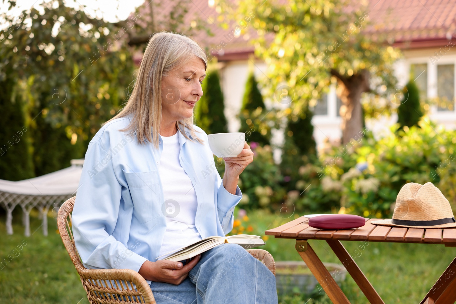 Photo of Senior woman with cup of drink and book in wicker armchair outdoors