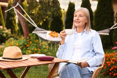 Photo of Senior woman with cup of drink and book in wicker armchair outdoors