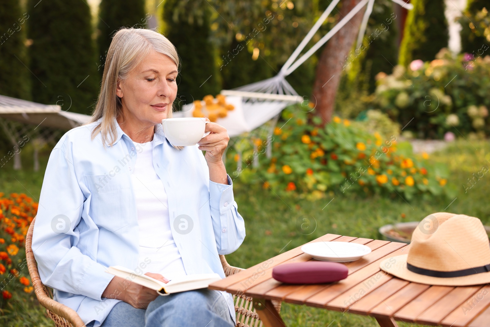 Photo of Senior woman with cup of drink and book in wicker armchair outdoors