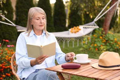 Photo of Senior woman with cup of drink and book in wicker armchair outdoors