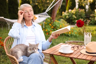 Photo of Senior woman with cat reading book at wooden table in garden