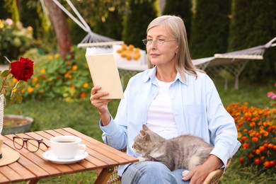 Photo of Senior woman with cat reading book at wooden table in garden