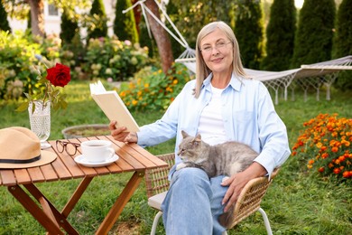 Photo of Senior woman with cat reading book at wooden table in garden