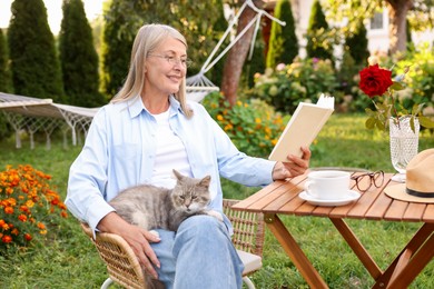 Photo of Senior woman with cat reading book at wooden table in garden