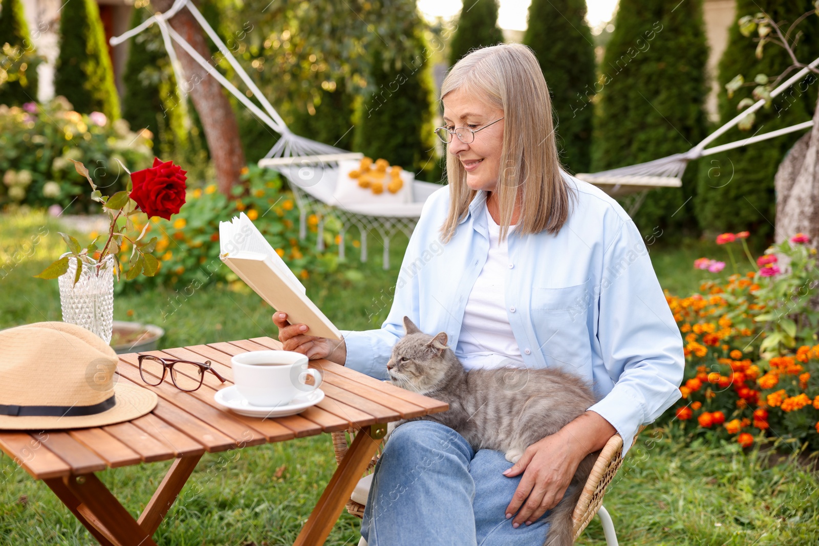 Photo of Senior woman with cat reading book at wooden table in garden