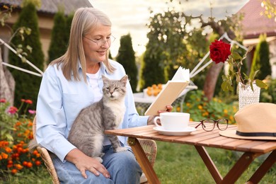 Photo of Senior woman with cat reading book at wooden table in garden