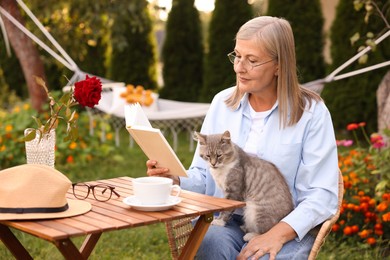 Photo of Senior woman with cat reading book at wooden table in garden