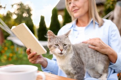 Photo of Senior woman stroking cat while reading book at table in garden, selective focus