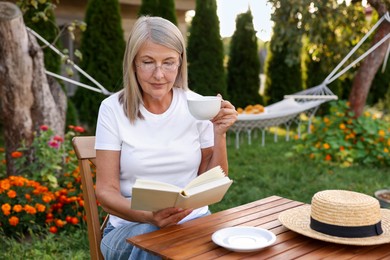 Photo of Senior woman with cup of drink reading book at wooden table in garden