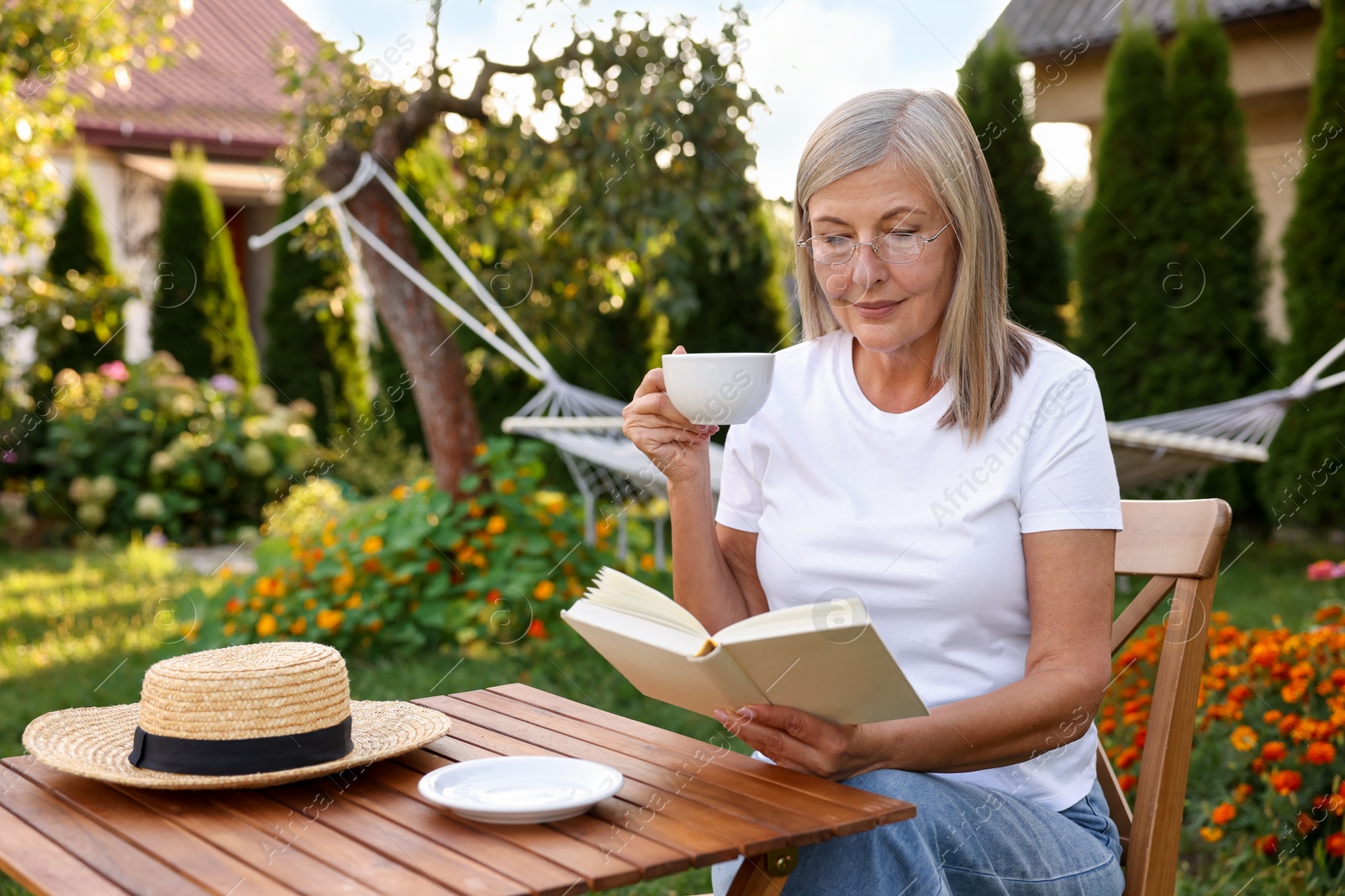 Photo of Senior woman with cup of drink reading book at wooden table in garden
