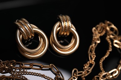 Photo of Stylish gold earrings and necklace on mirror surface against black background, closeup