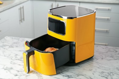Photo of Air fryer with cooked chicken drumsticks on white marble table in kitchen
