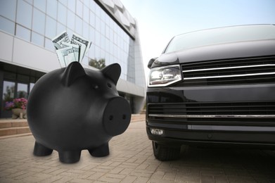 Image of Save money for car. Black piggy bank with dollars near automobile outdoors