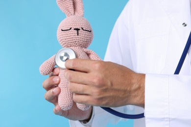 Photo of Pediatrician checking heartbeat of toy bunny on light blue background, closeup