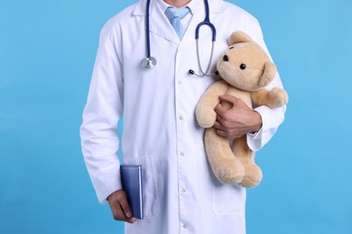Photo of Pediatrician with teddy bear and notebook on light blue background, closeup