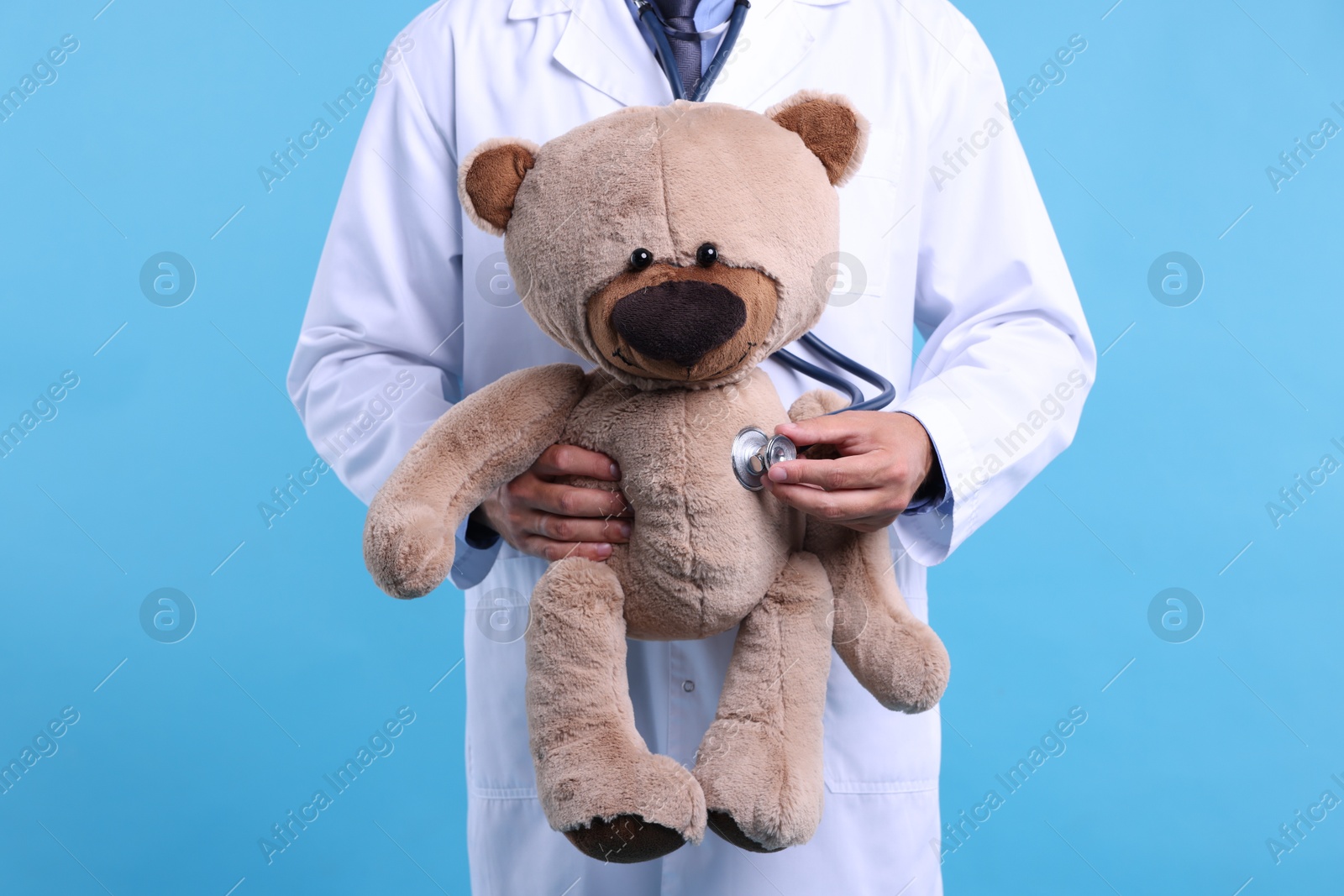 Photo of Pediatrician checking heartbeat of teddy bear on light blue background, closeup