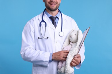 Photo of Pediatrician with toy bunny and clipboard on light blue background, closeup