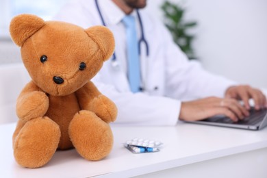 Photo of Pediatrician working on laptop in hospital, closeup. Teddy bear and pills on desk indoors