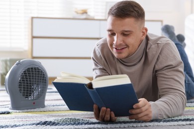 Photo of Young man reading book near electric heater at home