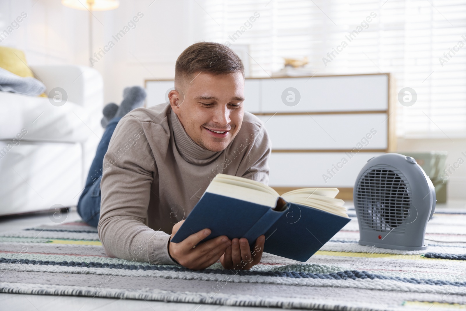 Photo of Young man reading book near electric heater at home